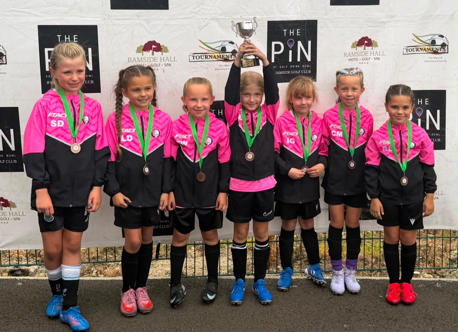 Young girls in pink FTACT club jackets celebrating with medals and a trophy
